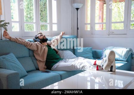 young relaxed man listening to music or podcast with headphones lying on the sofa at home. Stock Photo