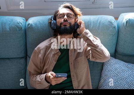 Young man wearing headphones listen podcast or music playing in smartphone sitting on sofa at home Stock Photo