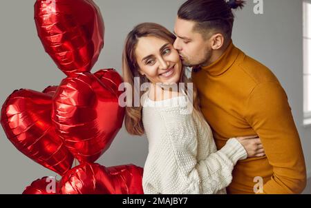 Man hugs his woman, kisses her on the cheek and gives her red heart shaped Valentine balloons Stock Photo