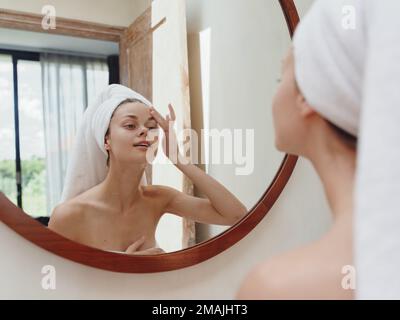 A beauty woman stands in front of a mirror after a shower in a towel on her head looks at her reflection and does a facial massage applies a day cream Stock Photo