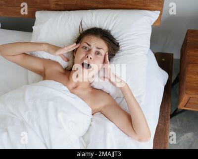 Woman lying on pillow under blanket in bed in bedroom, surprised face, open mouth, screaming Stock Photo
