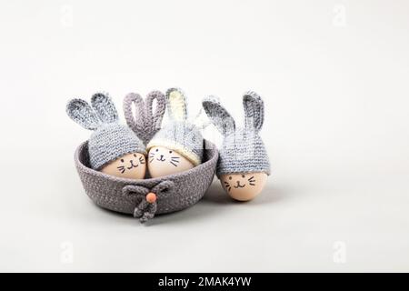 Three Easter eggs with funny faces and gray crocheted hats with bunny ears in a decorative basket on a gray background. Happy Easter concept. Greeting Stock Photo