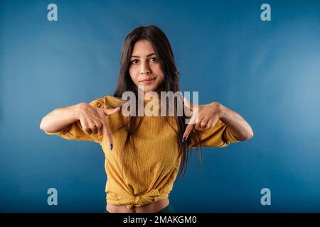 Young caucasian woman wearing casual top isolated over blue background pointing downward looks dissatisfied. Pointing downwards with index fingers. Stock Photo