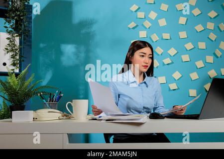 Confident Asian business woman doing paperwork while sitting at office desk. Focused entrepreneur using laptop with papers in hand preparing a report analyzing work results. Stock Photo