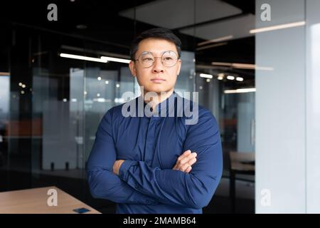 Asian serious and thinking man inside office looking out the window, businessman with crossed arms in shirt and sunglasses, boss thinking about an important decision. Stock Photo