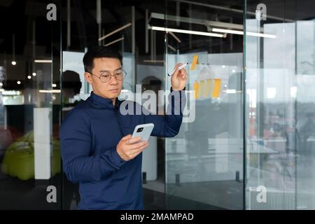 Asian man in thought writing down plans and strategy on colored paper on glass board, brainstorming in businessman at work. Stock Photo