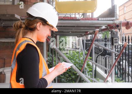 Smiley civil engineer lady carrying out an inspection and supervising a construction site building, wearing a hard hat and orange high visibility vest Stock Photo