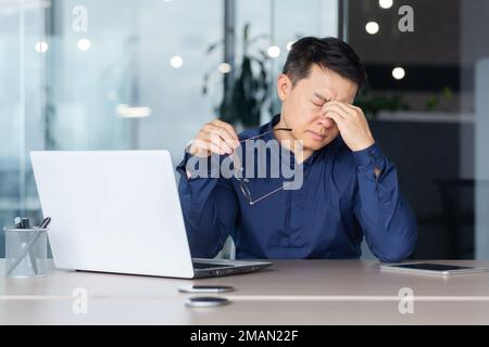 Asian worker tired at work has pain in eyes, businessman in glasses working late, using laptop at work inside office, serious and concentrated pensive. Stock Photo