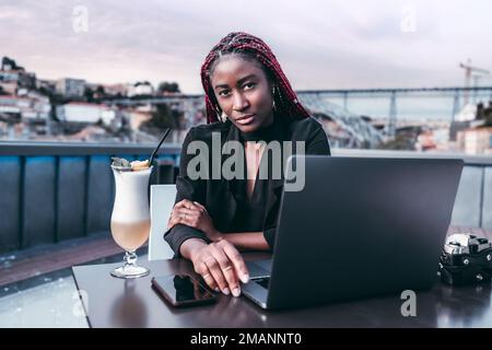 A mesmerizing black university student at a rooftop bar using her laptop while enjoying a cocktail on a cloudy day in the city of Porto on a defocused Stock Photo