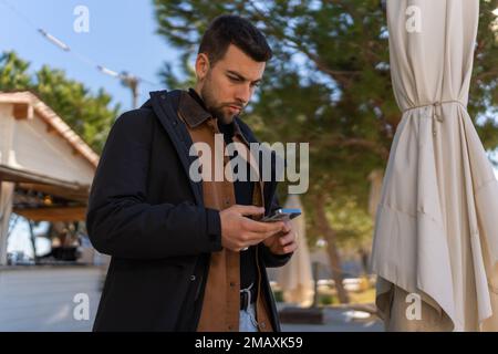 Low angle of concentrated male in outerwear standing in outdoor cafe and messaging on mobile phone Stock Photo