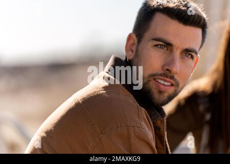 Young bearded guy with earring looking away thoughtfully while sitting on blurred background of street in sunny weather Stock Photo