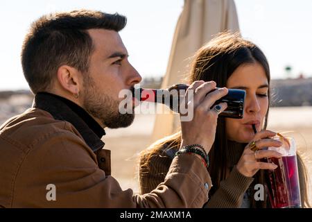 Young bearded male in outerwear drinking alcoholic beverage from glass bottle while woman sipping cold cocktail through straw on beach Stock Photo