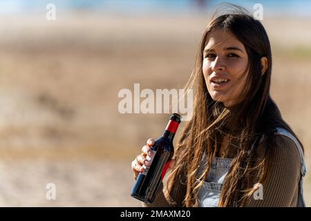 Young woman in casual clothing standing on blurred background of the beach during sunny and windy weather with a glass bottle of beer Stock Photo