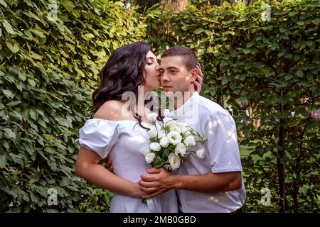 Girl in white dress gently hugs her boyfriend's neck and kisses him on the cheek. Newlyweds in the park. Portrait Stock Photo