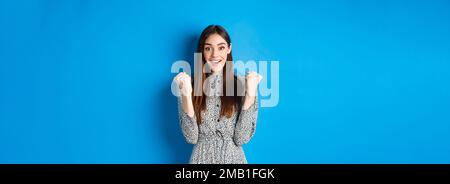 Excited woman looking relieved and hopeful, making fist pump and smiling, winning prize, celebrating achievement or victory, standing on blue Stock Photo