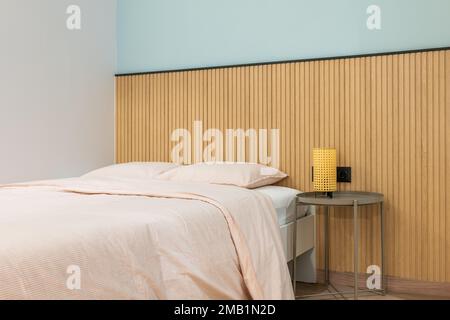 Double bed with pillows is made up with large warm duvet and linens in pleasant beige color. On table is lamp with yellow frame for soft warm light at Stock Photo