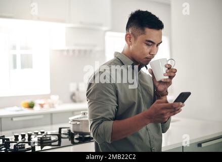 Doing some morning browsing. a young man using a cellphone while drinking coffee in the kitchen at home. Stock Photo