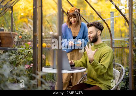 Couple works remotely on laptop in glasshouse at backyard Stock Photo