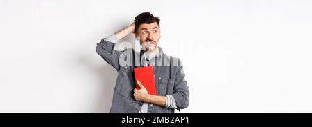 Confused guy scratch head and look aside at logo puzzled, holding red diary or planner, standing against white background Stock Photo