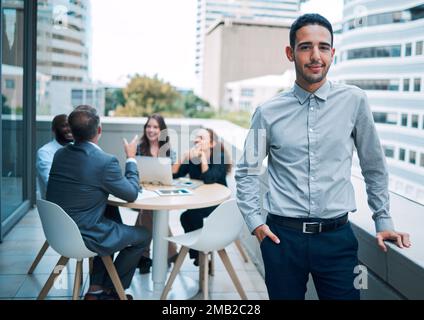 A leader is one who knows, goes and shows the way. Portrait of a confident young businessman standing on the balcony of an office with his colleagues Stock Photo