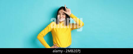 Sassy asian girl mocking lost team, showing loser sign on forehead and smiling pleased, being a winner, standing over blue background. Stock Photo