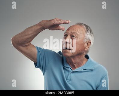 What is that over there. an older man with his hand covering his face looking off into the distance in a studio against a grey background. Stock Photo