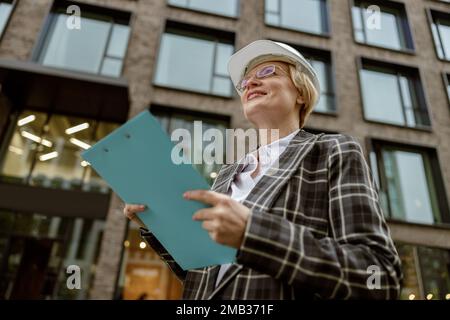 Senior female engineer in suit with white safety helmet holding tablet on city building background Stock Photo