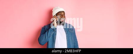 Hipster Black man talking on phone, looking left and having mobile conversation, standing over pink background Stock Photo