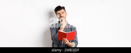 Thoughtful young man plan his schedule, looking aside pensive, holding opened planner or journal, standing on white background Stock Photo