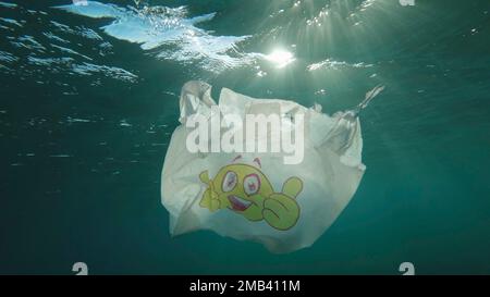 Plastic pollution, used plastic bag with smiley face slowly drifting underwater in sun rays on dawn. Low-angle shot, Backlight (Contre-jour) . Stock Photo