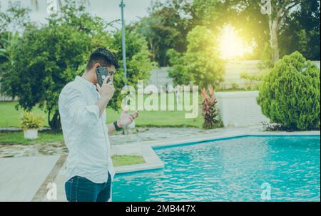 Attractive guy standing talking on the phone near the pool edge. Handsome man calling on the phone by a swimming pool, Young man standing calling on Stock Photo