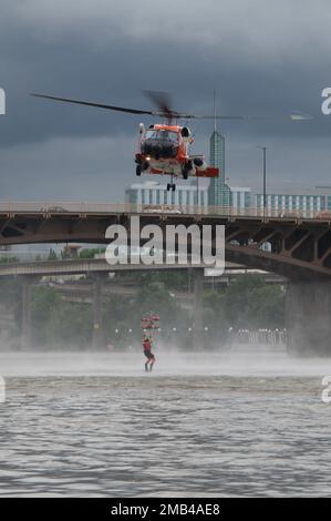 A Coast Guard aviation survival technician holds on to the basket  pulled out of the water by an MH-60 Jayhawk helicopter crew on the Willamette River in Portland, Oregon, June 11, 2022. The AST guided the basket deployment for the demonstration. Stock Photo