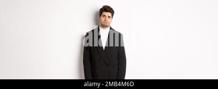 Image of gloomy and sad handsome male model in black suit, frowning and looking tired at camera, standing against white background. Stock Photo