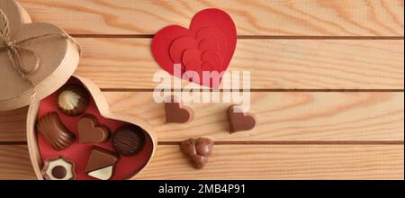 Box with assorted chocolates and heart-shaped red cut-outs on wooden slatted table. Top view. Stock Photo