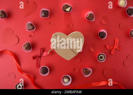 Chocolates on paper mold on a red table decorated with hearts and a heart-shaped brown gift box in the center. Top view. Stock Photo