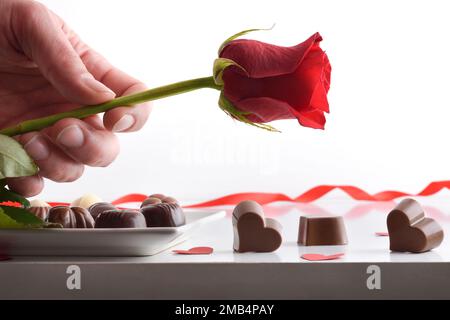 Hand offering a rose and assortment of chocolates served on a plate on a white table with several heart-shaped chocolates and a white isolated backgro Stock Photo