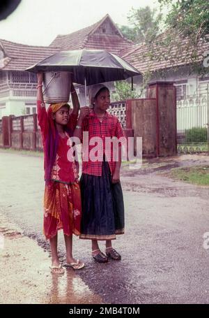 Two girl children with umbrella on a rainy day in Kottayam, Kerala, India, Asia. One girl with a bucket over her head. Monsoon, Umbrella, Street Stock Photo