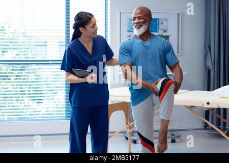 Smiling diverse female physiotherapist and senior male patient holding leg in therapy, copy space Stock Photo