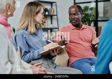 African American man telling about his problems to specialist while she making notes in notepad during session in the room Stock Photo