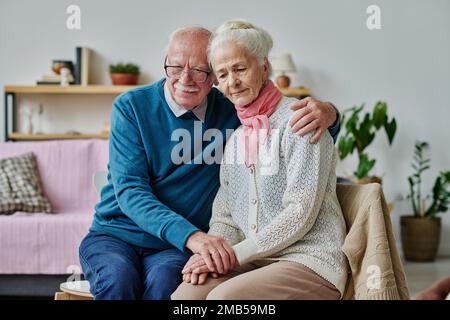 Senior man embracing his wife in depression and supporting her while they sitting on chairs in the room Stock Photo
