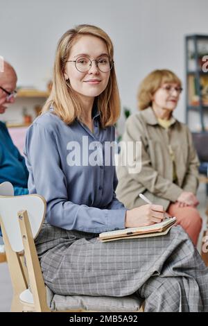 Portrait of young woman looking at camera while making notes in her notepad during studying with other people in background Stock Photo