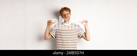 Confident young nerdy guy in glasses with red hair, pointing at himself and looking like professional, white background Stock Photo