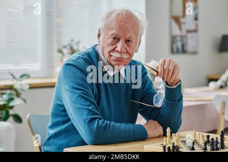 Portrait of senior man with grey hair looking at camera while playing chess at table Stock Photo