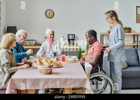 Senior people sitting at table and eating with caregiver helping man with disability in nursing home Stock Photo