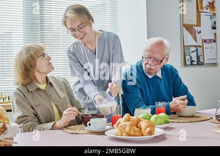 Young volunteer caring about senior people during lunch, she pouring milk in bowl during breakfast Stock Photo