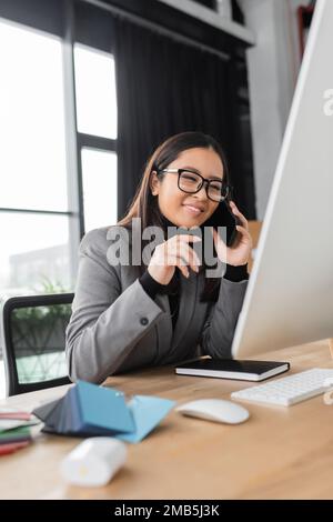 Smiling asian interior designer talking on smartphone near computer and color samples,stock image Stock Photo