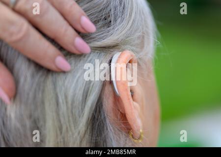 Elderly gray haired hard of hearing lady in her 80's brushes her hair to the side to show her hearing aid in her ear Stock Photo