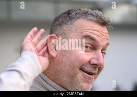 Middle age grey-haired hard of hearing man in his fifties or sixties holds his hand behind his ear in which he has a hearing aid Stock Photo
