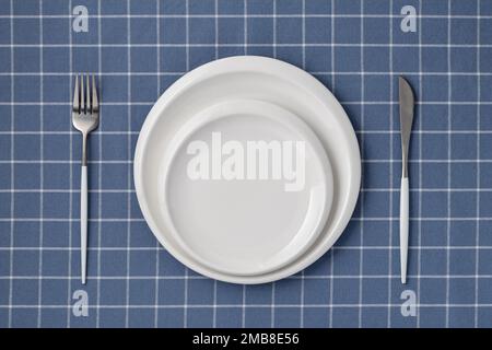 White empty plate and cutlery on a blue tablecloth with checkered pattern, table top view, above. Flat lay design. Ceramic utensils, serving. Tablewar Stock Photo