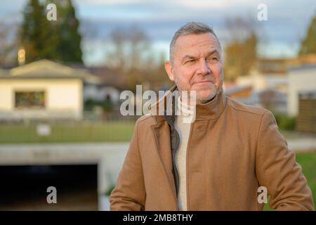 Close-up of a middle-aged man in a brown coat looking thoughtfully to the side against an urban background Stock Photo